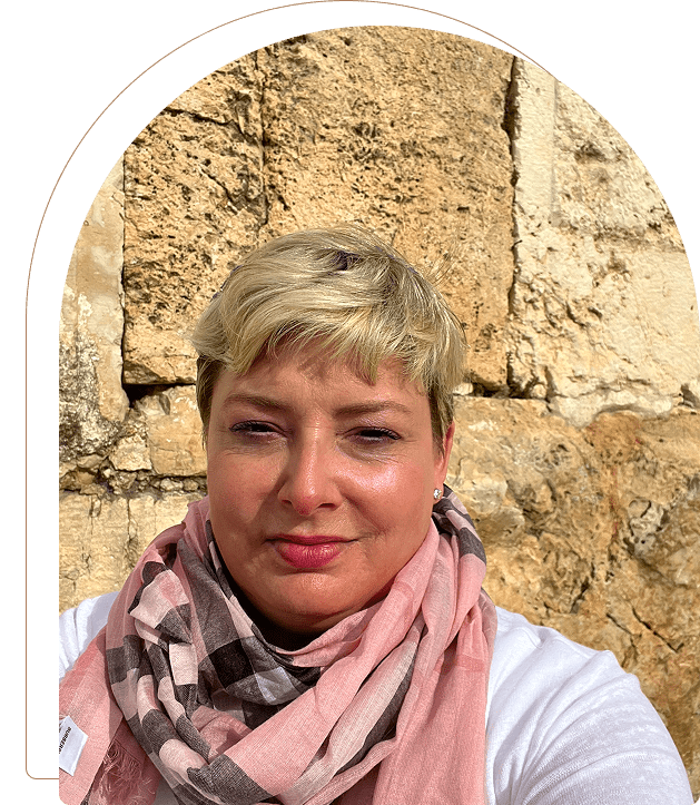 Woman with short blonde hair and a pink scarf in front of a stone wall.